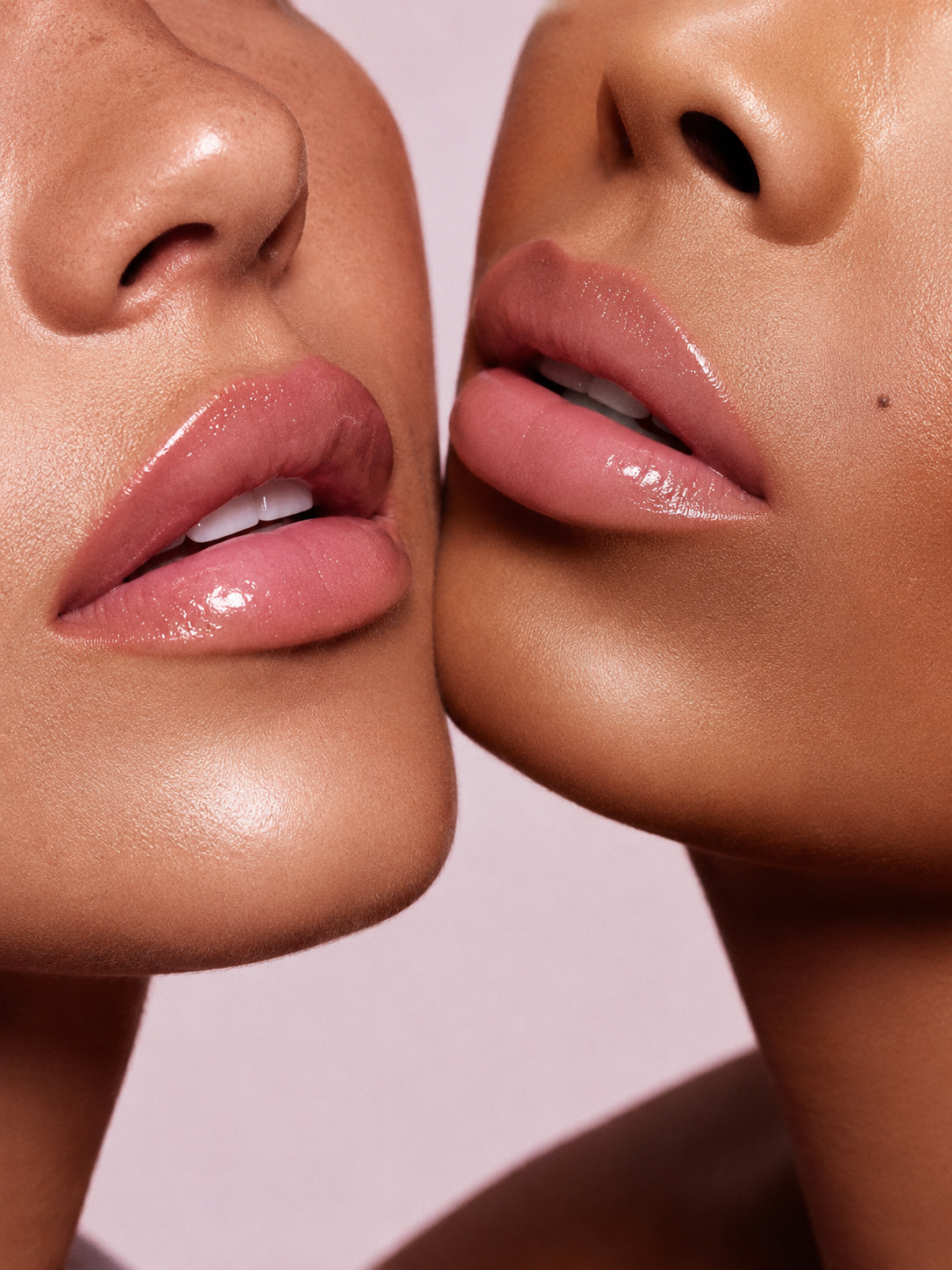 Deux femmes aux carnations différentes portant un gloss rose nude, photographiées en gros plan sur fond rose poudré.